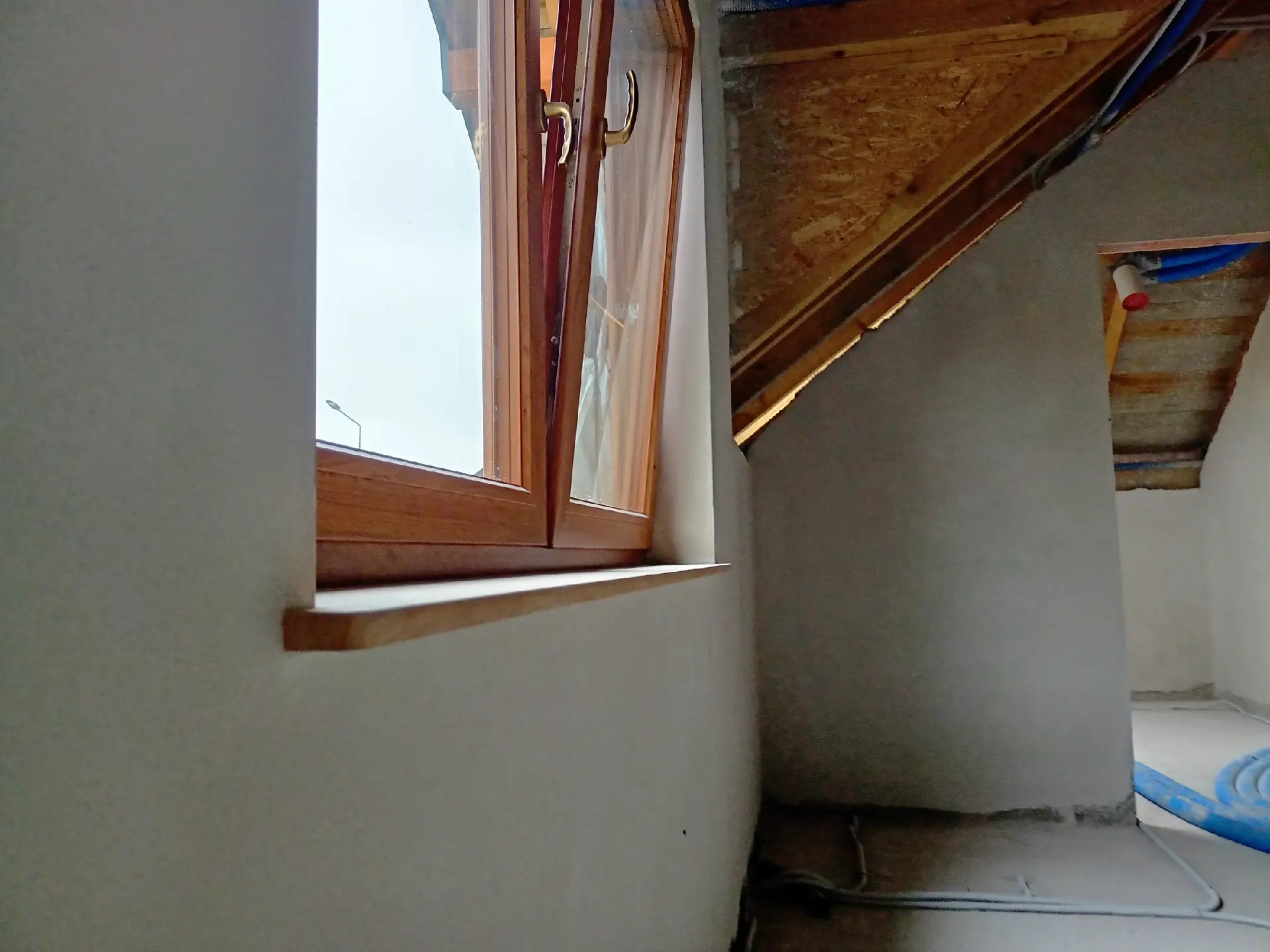 Poddasze w trakcie prac wykończeniowych: drewniane okno dachowe z otwartym skrzydłem, surowy tynk na ścianach, widoczne instalacje pod stropem.