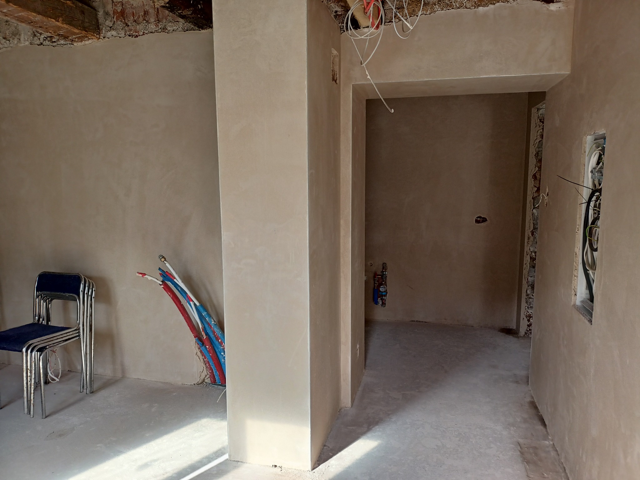 Wnętrze w trakcie remontu: świeżo otynkowane ściany w odcieniach beżu, widoczne wystające rury instalacyjne w kolorach czerwonym i niebieskim, oraz stos krzeseł z niebieskimi siedziskami.
