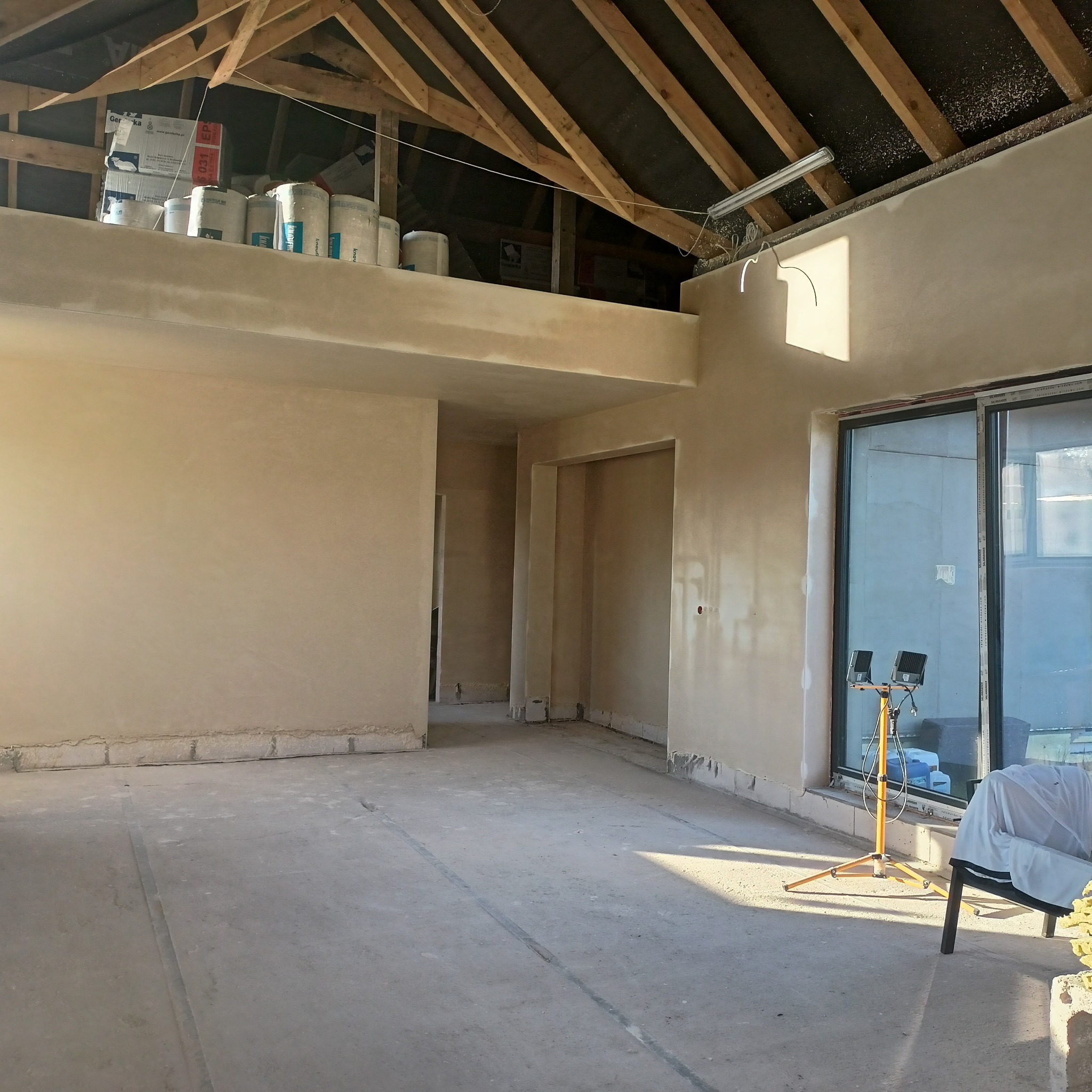Wnętrze w stanie surowym po nałożeniu gładzi, widoczne ślady po pracach, reflektor budowlany i materiały budowlane na antresoli pod drewnianą konstrukcją dachu.