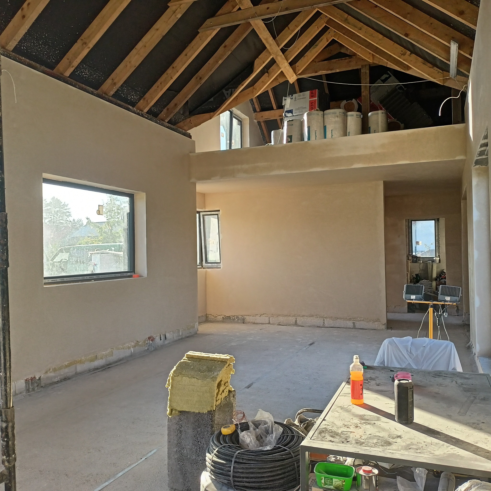 Surowe, jasne wnętrze w trakcie prac wykończeniowych, ściany pokryte świeżym tynkiem, widoczne drewniane elementy konstrukcji dachu, izolacja na podłodze i narzędzia na stole roboczym.