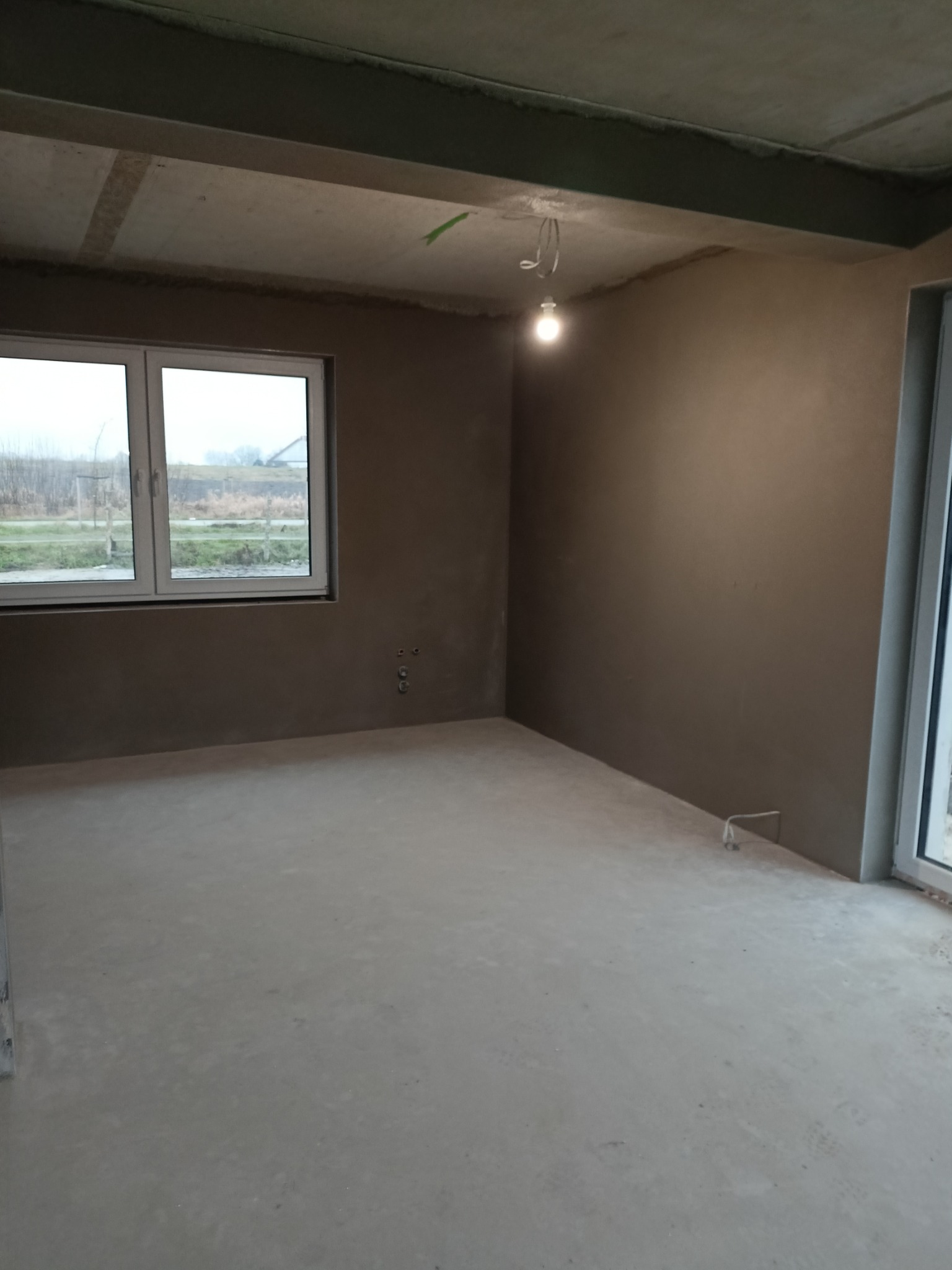 Surowe wnętrze po nałożeniu gładzi gipsowej na ściany, widoczne przyłącza instalacji, surowy betonowy strop i prowizoryczne oświetlenie żarówką