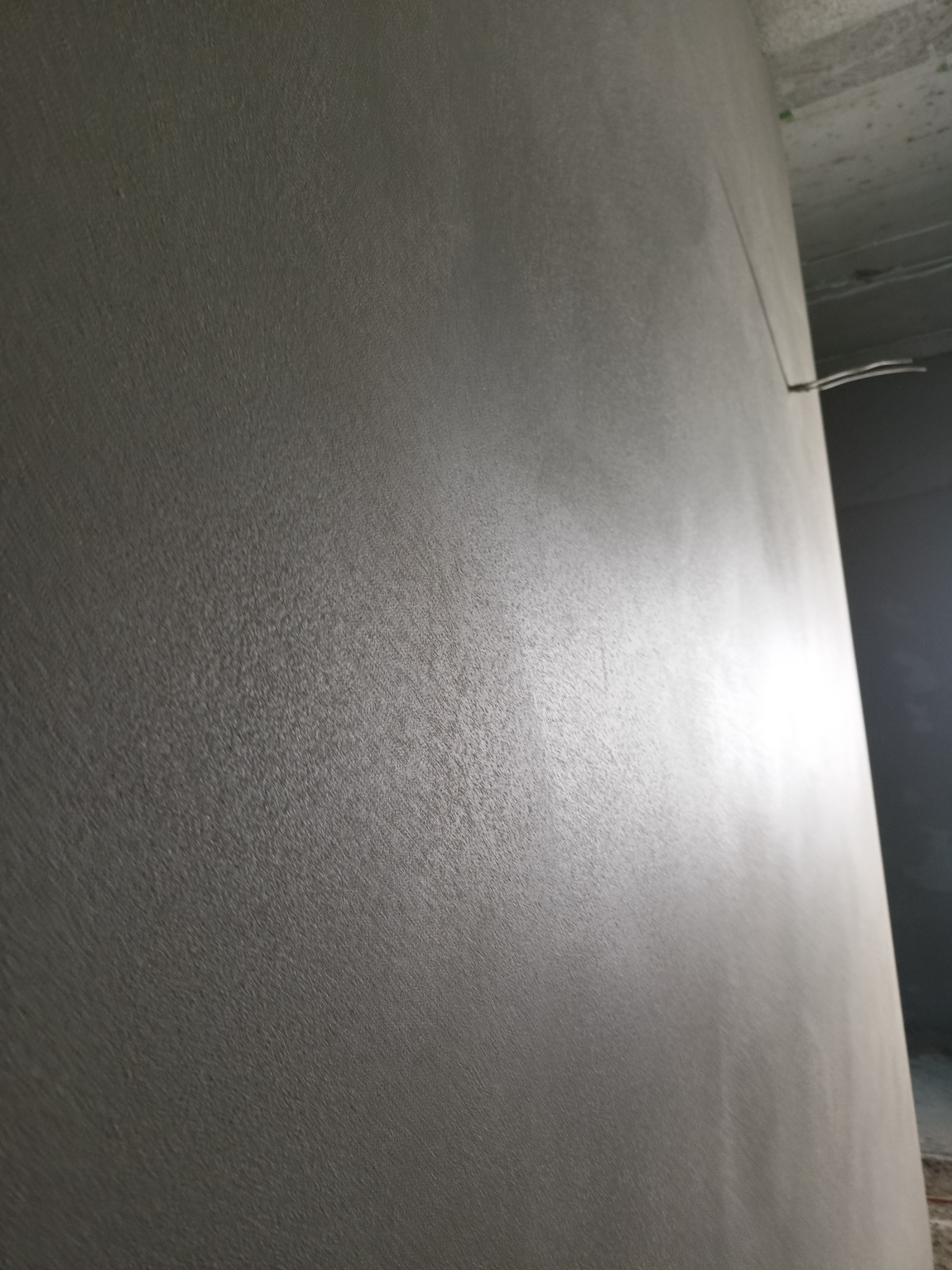 Świeżo nałożona, szara warstwa tynku na ścianie wewnętrznej z widocznymi przewodami elektrycznymi wystającymi z górnej krawędzi ściany.