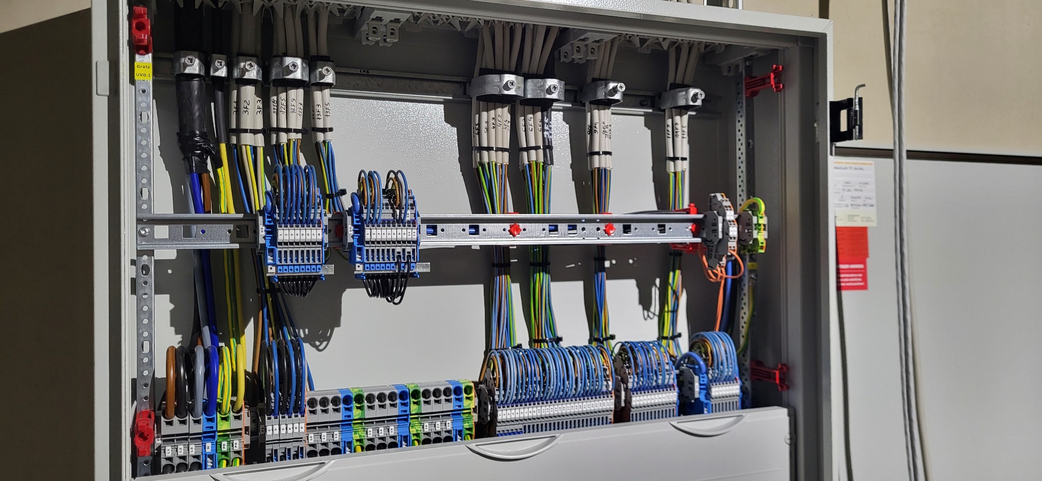 Rozdzielnia elektryczna z precyzyjnie ułożonymi przewodami oznaczonymi etykietami, zamontowana w metalowej obudowie. Widoczne złącza i listwy montażowe.