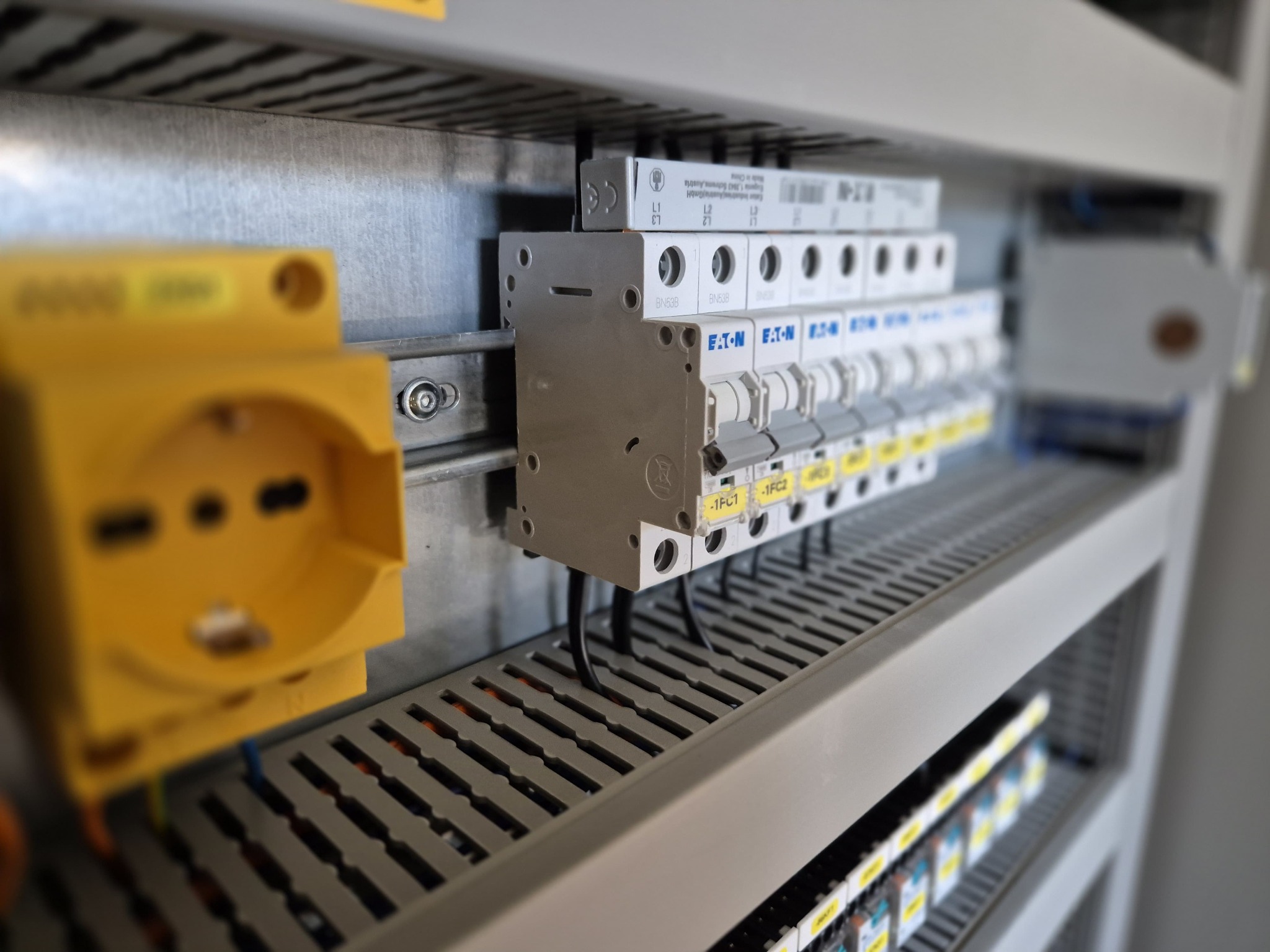 Szczegółowe ujęcie rozdzielnicy elektrycznej z widocznymi bezpiecznikami EATON, żółtym gniazdkiem i przewodami w szarej obudowie. Widok z boku, częściowo rozmyte tło.