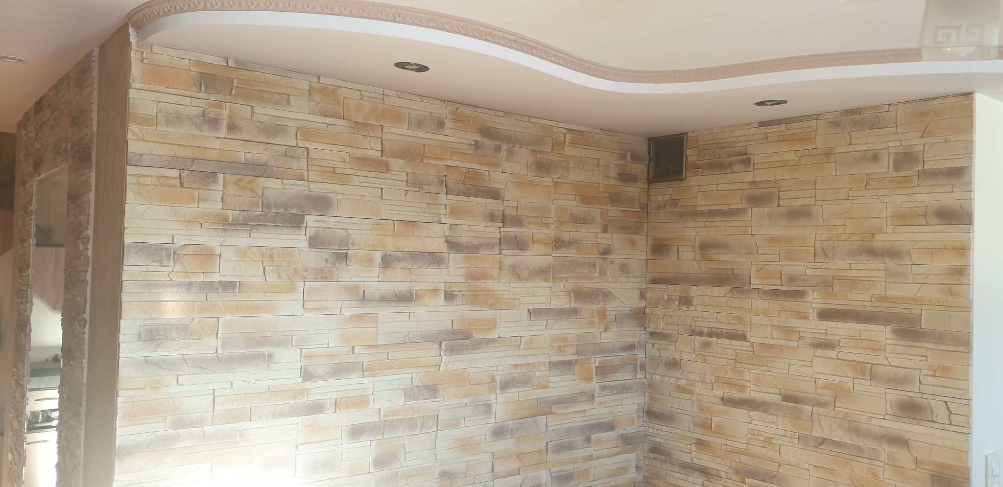 Ściana wykończona dekoracyjną okładziną imitującą kamień w odcieniach beżu i brązu, z widocznym fragmentem sufitu z ozdobnym gzymsem.