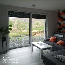 F.H.U. DARIUSZ RAKOWSKI - Wnętrze salonu z dużym oknem przesuwnym z roletami zewnętrznymi, szary narożnik z pomarańczowymi poduszkami, biały stolik kawowy i podłoga imitująca drewno, za oknem widok na teren w trakcie prac...