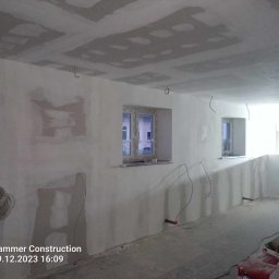 F.H.U. DARIUSZ RAKOWSKI - Wnętrze w trakcie remontu: ściany z częściowo nałożoną gładzią, widoczne kable elektryczne, sufit z płyt kartonowo-gipsowych, dwa okna z widokiem na budynek na zewnątrz, materiały budowlane w rogu...