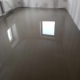 F.H.U. DARIUSZ RAKOWSKI - Świeżo wylana, mokra wylewka samopoziomująca w pokoju z oknami, grzejnikiem owiniętym folią i ścianami przygotowanymi do dalszych prac wykończeniowych.