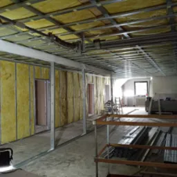 Wnętrze w trakcie prac remontowych, widoczne metalowe profile konstrukcyjne ścian działowych z izolacją z wełny mineralnej, sufit podwieszany w trakcie montażu, oświetlenie robocze, oraz materiały...