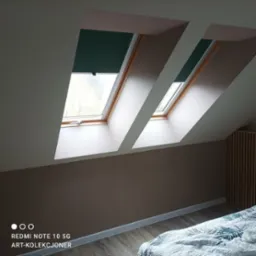 Pokój na poddaszu z dwoma oknami dachowymi z roletami, ściany pomalowane na brązowo, listwy przypodłogowe, drewniana podłoga i fragment kołdry na łóżku.