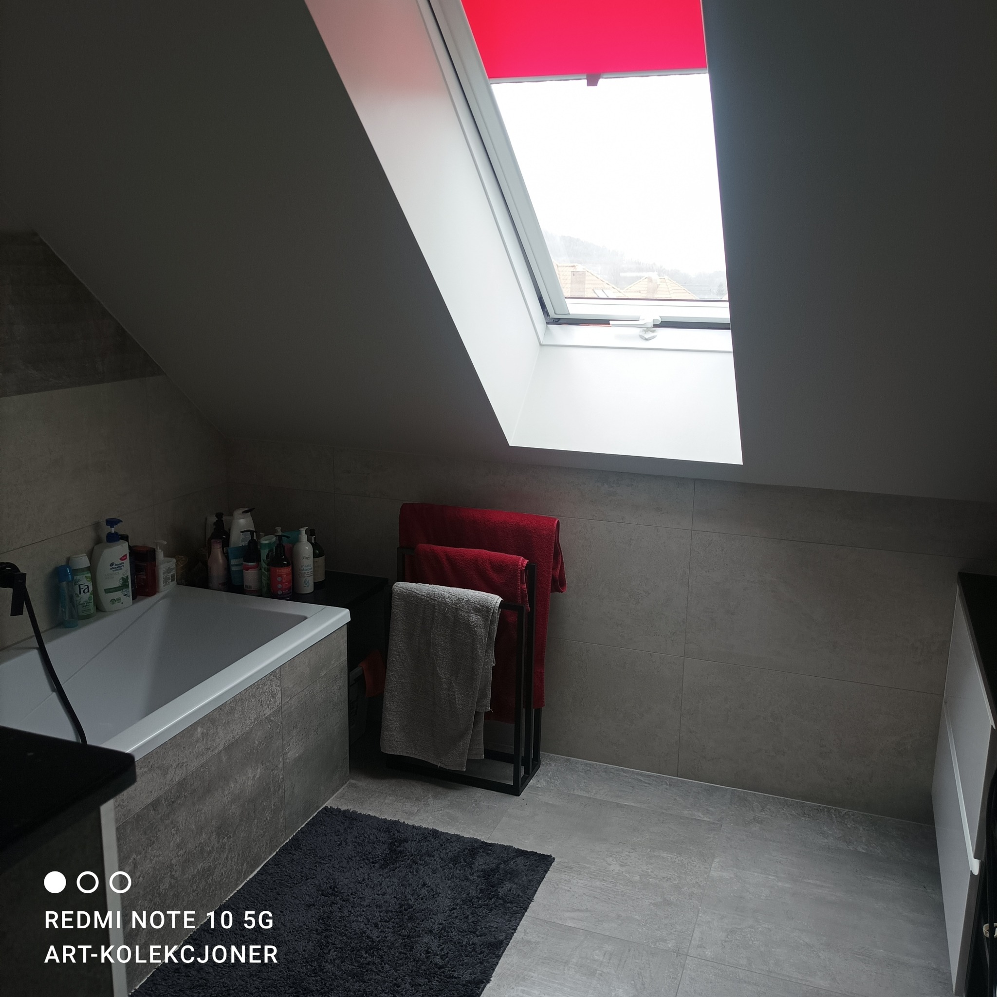 Szara łazienka na poddaszu z oknem dachowym z czerwoną roletą, płytkami imitującymi beton, wanną, czarnym dywanikiem i wieszakiem na ręczniki z czerwonymi i szarymi ręcznikami.