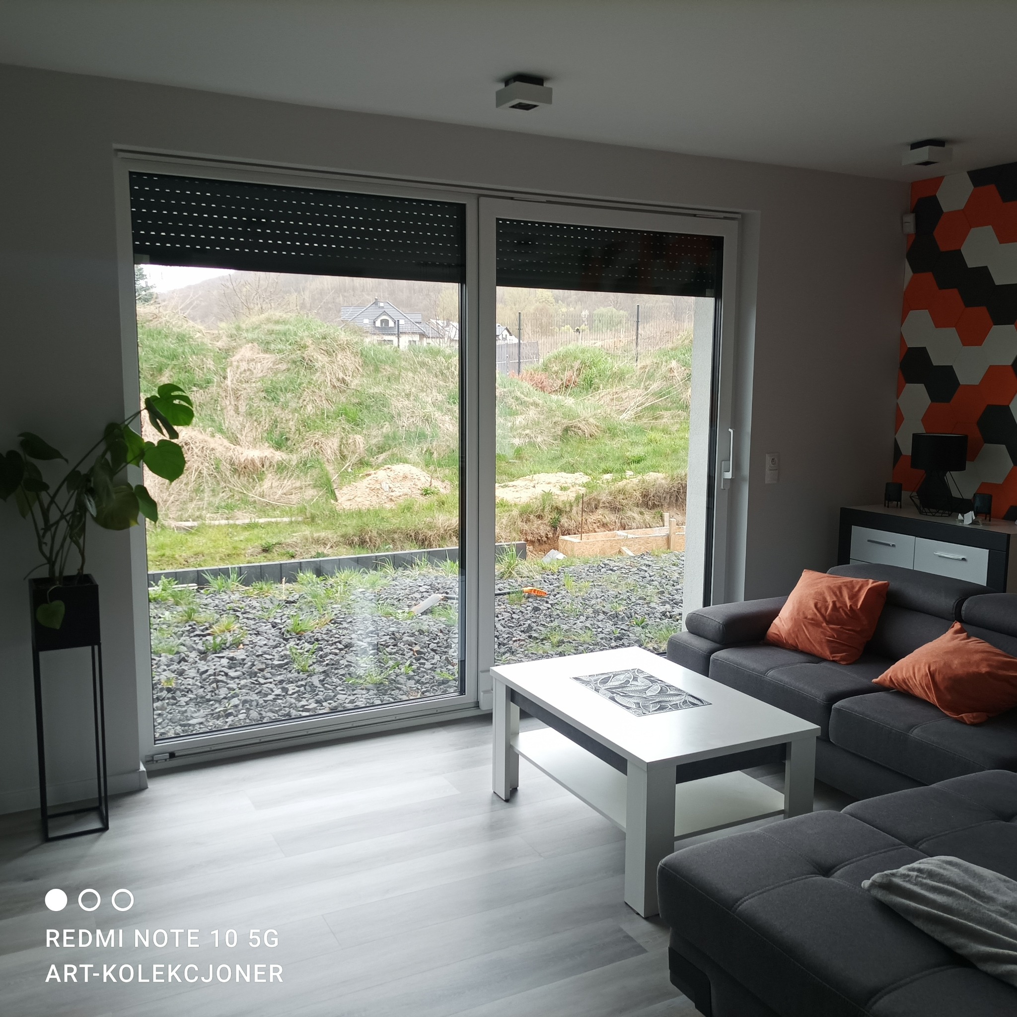 Wnętrze salonu z dużym oknem przesuwnym z roletami zewnętrznymi, szary narożnik z pomarańczowymi poduszkami, biały stolik kawowy i podłoga imitująca drewno, za oknem widok na teren w trakcie prac...