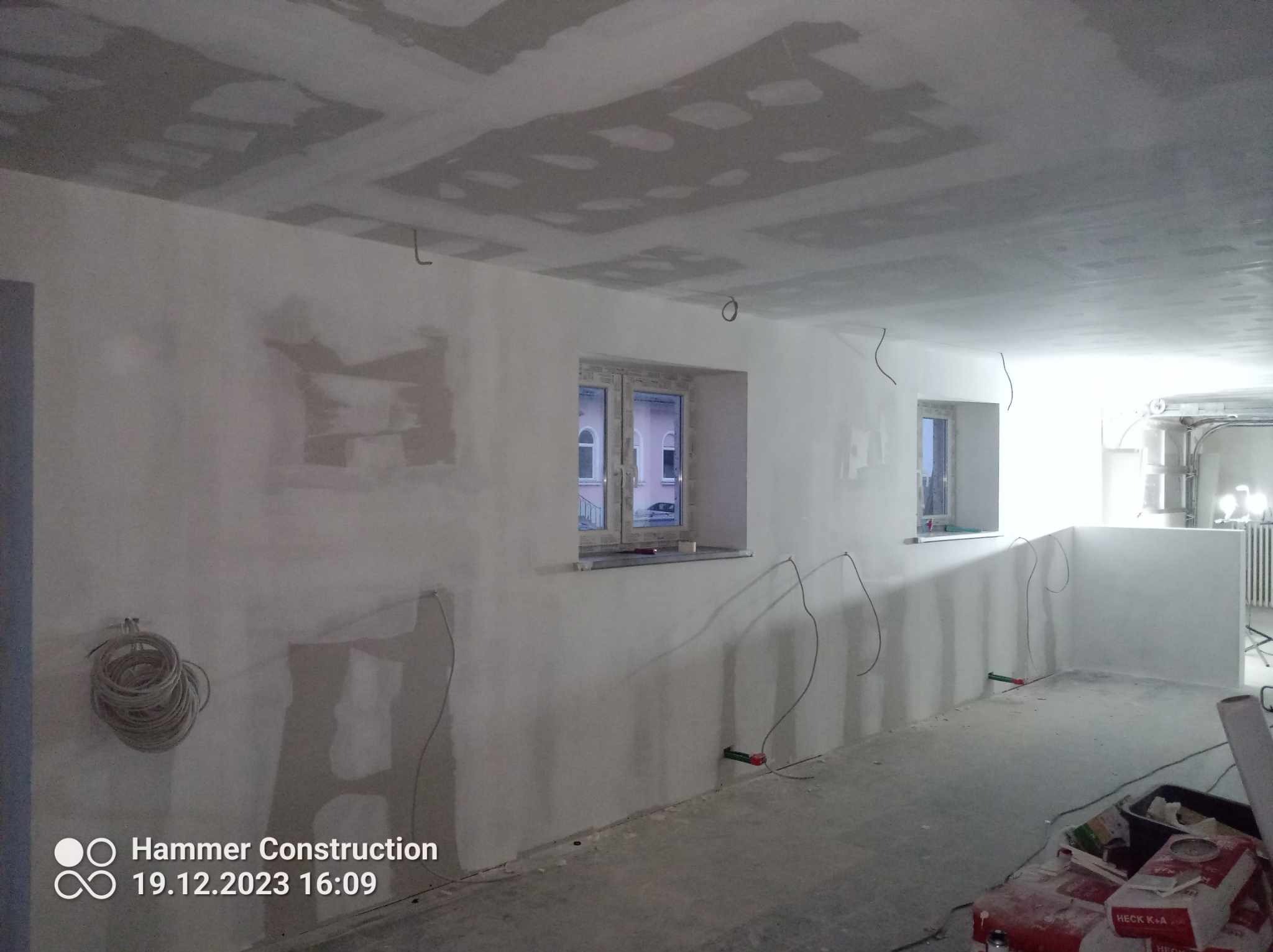 Wnętrze w trakcie remontu: ściany z częściowo nałożoną gładzią, widoczne kable elektryczne, sufit z płyt kartonowo-gipsowych, dwa okna z widokiem na budynek na zewnątrz, materiały budowlane w rogu...
