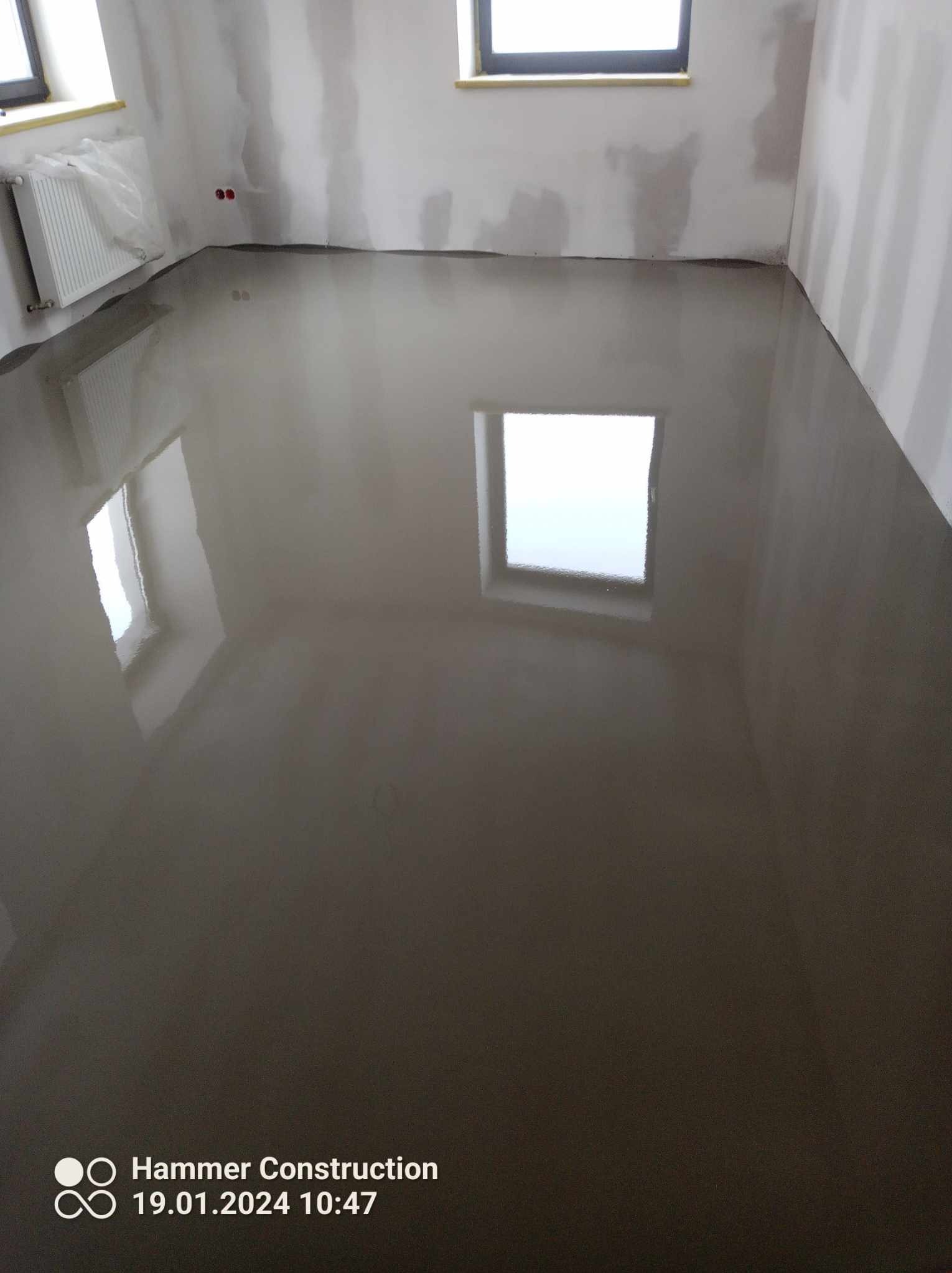 Świeżo wylana, mokra wylewka samopoziomująca w pokoju z oknami, grzejnikiem owiniętym folią i ścianami przygotowanymi do dalszych prac wykończeniowych.