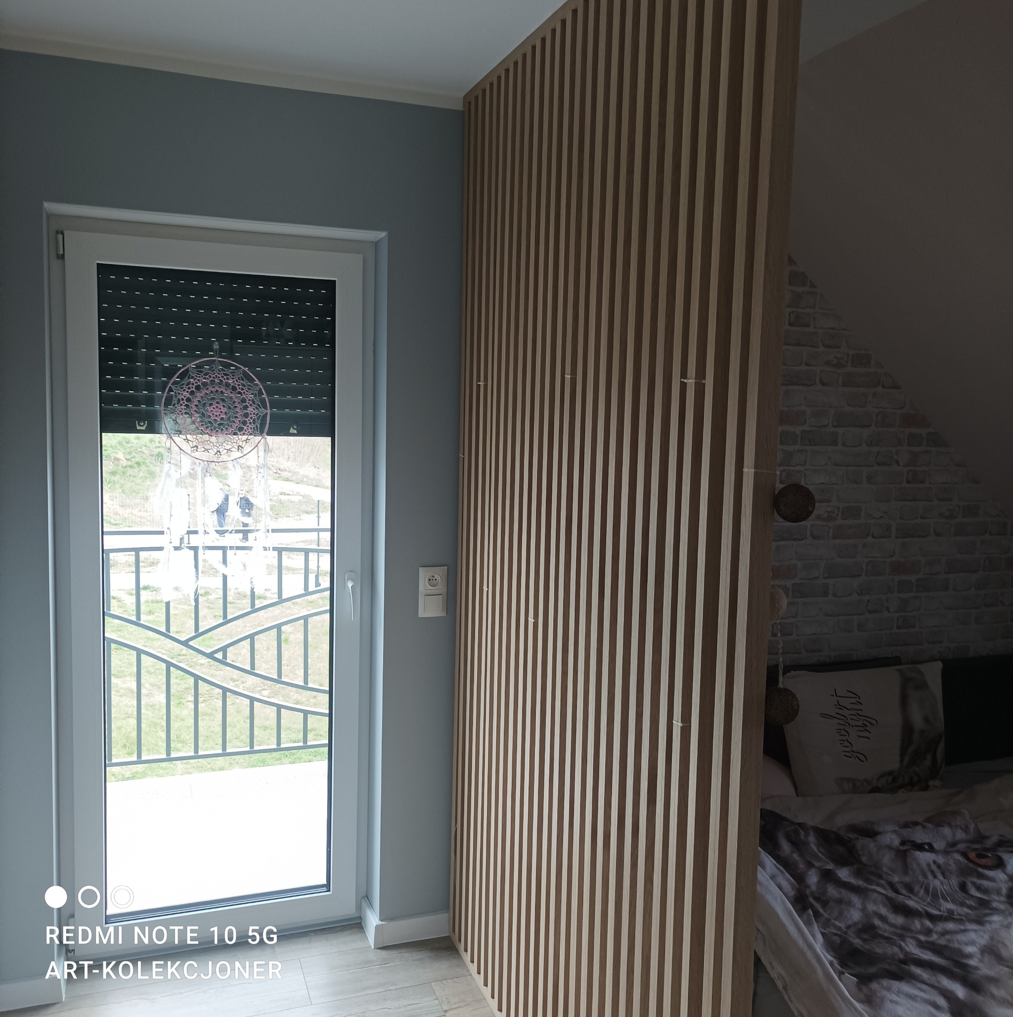 Drewniane lamele oddzielające przestrzeń sypialnianą od okna z widokiem na zewnątrz, w tle ściana z cegły i pościel z motywem wilka.