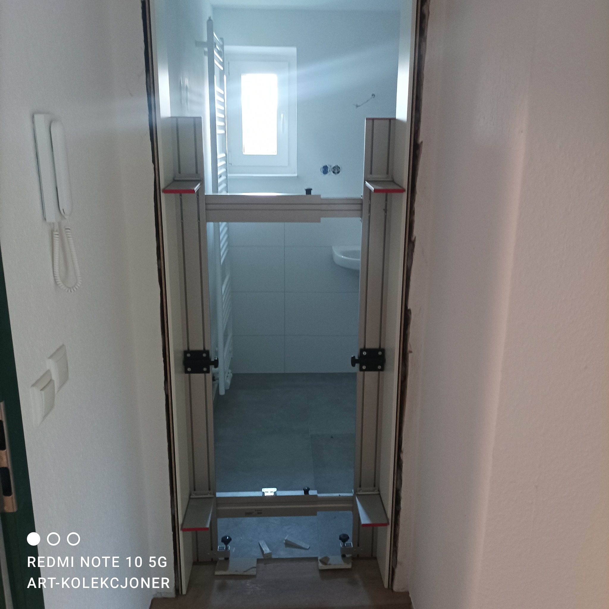 Widok na łazienkę przez zamontowaną ościeżnicę regulowaną z aluminium, widoczne białe płytki, umywalka i grzejnik drabinkowy.