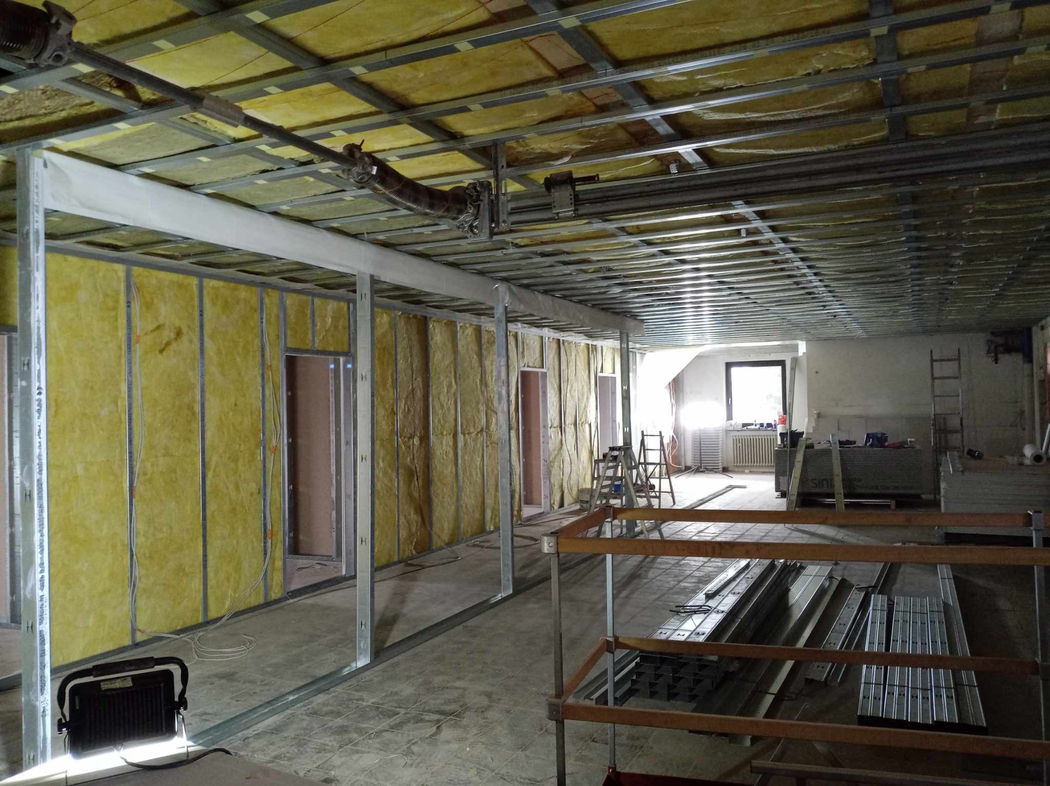 Wnętrze w trakcie prac remontowych, widoczne metalowe profile konstrukcyjne ścian działowych z izolacją z wełny mineralnej, sufit podwieszany w trakcie montażu, oświetlenie robocze, oraz materiały...