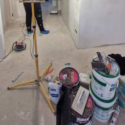 REM-ART ARTUR AFTYKA - Przygotowania do prac wykończeniowych: widoczne wiaderka z masą szpachlową, worek z gipsem szpachlowym Knauf Uniflott, szpachelki, paca, przedłużacz elektryczny oraz lampa budowlana na trójnogu,...