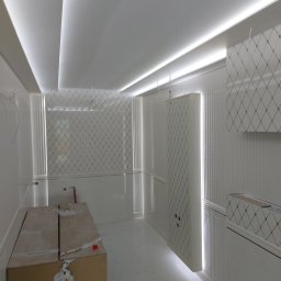 REM-ART ARTUR AFTYKA - Wnętrze kuchni w trakcie remontu, widoczne białe panele ścienne z wzorem rombów, podświetlany sufit i kartony z materiałami budowlanymi na podłodze.