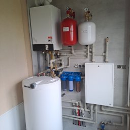 Tomasz Kępa - Kompletna instalacja grzewcza i wodna z białym kotłem Viessmann, dużym zasobnikiem, filtrami wody oraz czerwonym i białym naczyniem wzbiorczym, zamontowana na szarej ścianie.