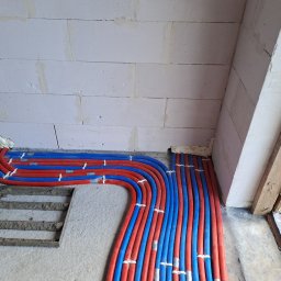 Tomasz Kępa '' BORGOSZ ENERGY '' - Precyzyjnie ułożone czerwone i niebieskie rury instalacji grzewczej podłogowej na betonowej posadzce, zabezpieczone opaskami, przy ścianie z białych bloczków.