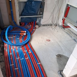 Tomasz Kępa '' BORGOSZ ENERGY '' - Gęsto ułożone czerwone i niebieskie rury systemu grzewczego na surowej betonowej posadzce, mocowane opaskami. W tle białe przewody i niebieska rura ścienna.