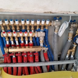 Tomasz Kępa '' BORGOSZ ENERGY '' - Mosiężny rozdzielacz ogrzewania podłogowego z niebieskimi i czerwonymi rurami w izolacji, zamontowany w szarej skrzynce ściennej, z widocznym licznikiem wody i żółtą izolacją u dołu.
