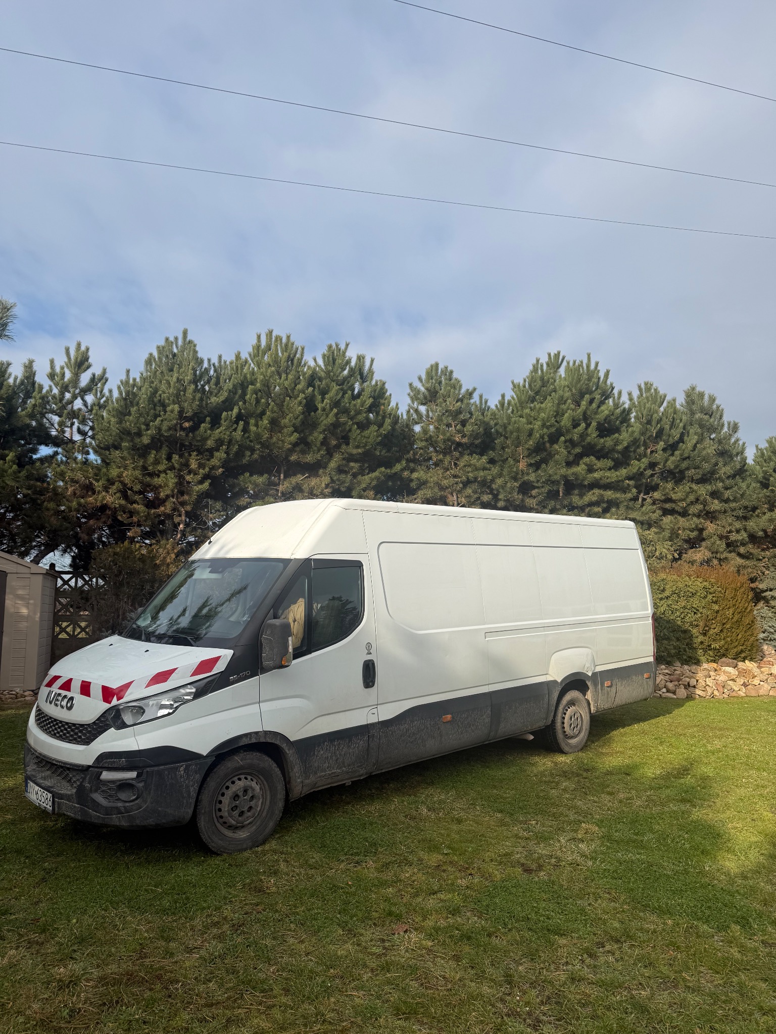 Biały bus dostawczy Iveco na trawniku, z widocznymi oznaczeniami ostrzegawczymi na masce, sugerujący transport lub przeprowadzkę. Tło stanowią drzewa i krzewy.