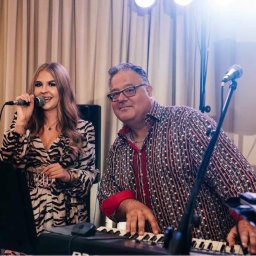 Gajewski Band - Wokalistka w sukience ze wzorem zebry śpiewa do mikrofonu, obok pianista w koszuli w kropki gra na keyboardzie, za nimi kremowe zasłony i reflektory.