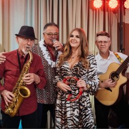 Gajewski Band - Zespół muzyczny: saksofonista w kapeluszu, wokalista z mikrofonem w hawajskim naszyjniku, kobieta z tamburynem w sukience w zebrę i gitarzysta w białej koszuli, oświetlenie sceniczne w tle.
