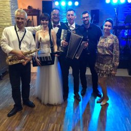 Gajewski Band - Zespół muzyczny w składzie: saksofonista, klawiszowiec, akordeonista i wokalistka, pozujący na tle dekoracji sali weselnej z oświetleniem scenicznym.