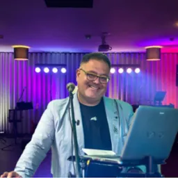 Uśmiechnięty mężczyzna w okularach i jasnej marynarce, stoi za mikrofonem i laptopem marki Sony, na tle sceny z nagłośnieniem, oświetleniem i instrumentami muzycznymi.