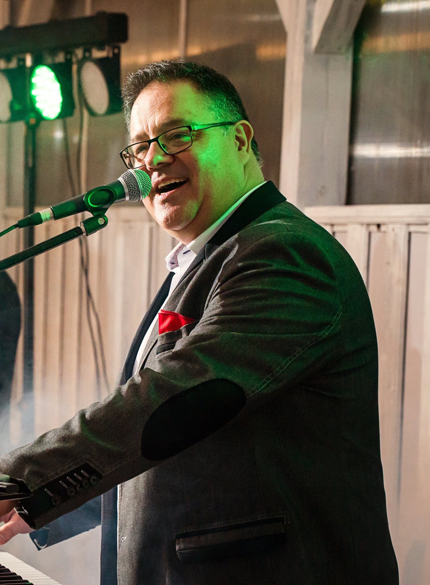 Mężczyzna w okularach i marynarce z czerwoną poszetką śpiewa do mikrofonu, grając na keyboardzie pod zielonym oświetleniem scenicznym.