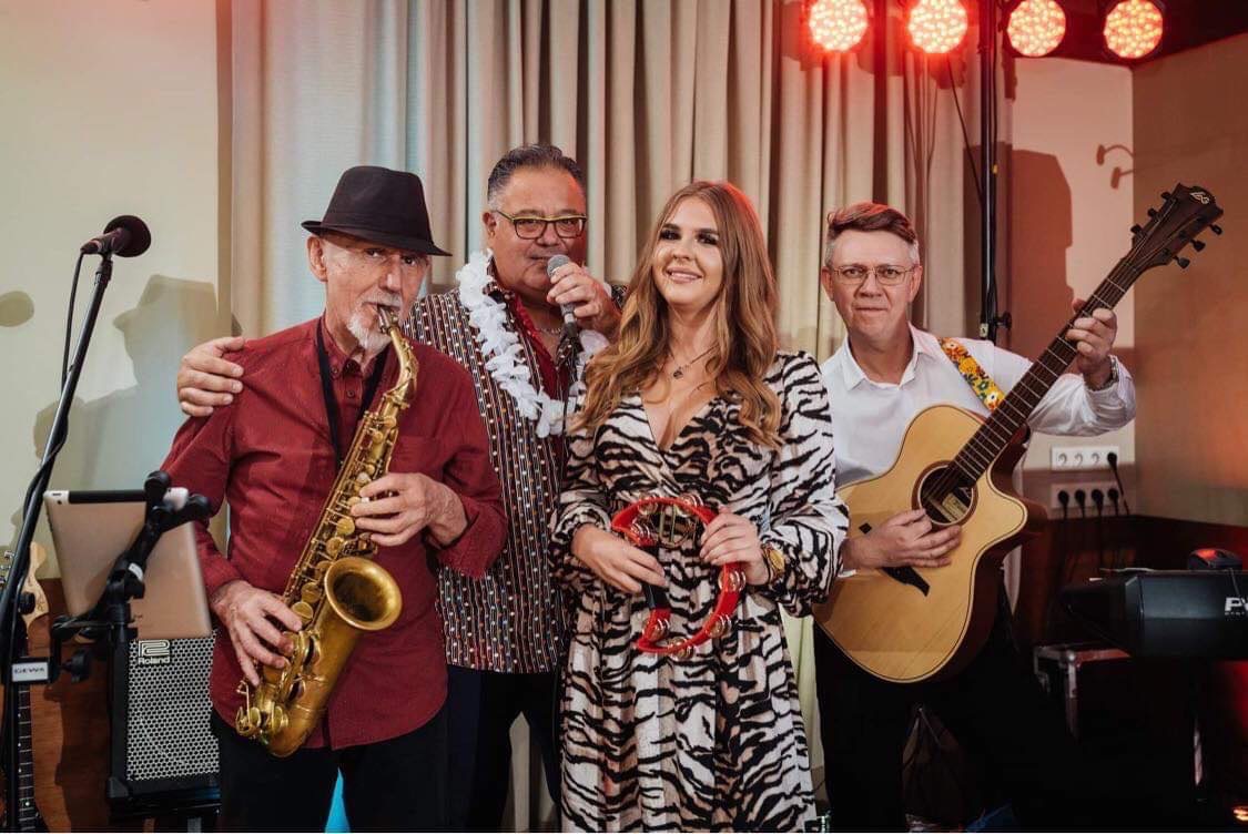 Zespół muzyczny w trakcie występu: saksofonista w kapeluszu, wokalista z mikrofonem w hawajskim naszyjniku, kobieta z tamburynem w sukience w zebrę i gitarzysta z kolorowym paskiem na tle kotary...