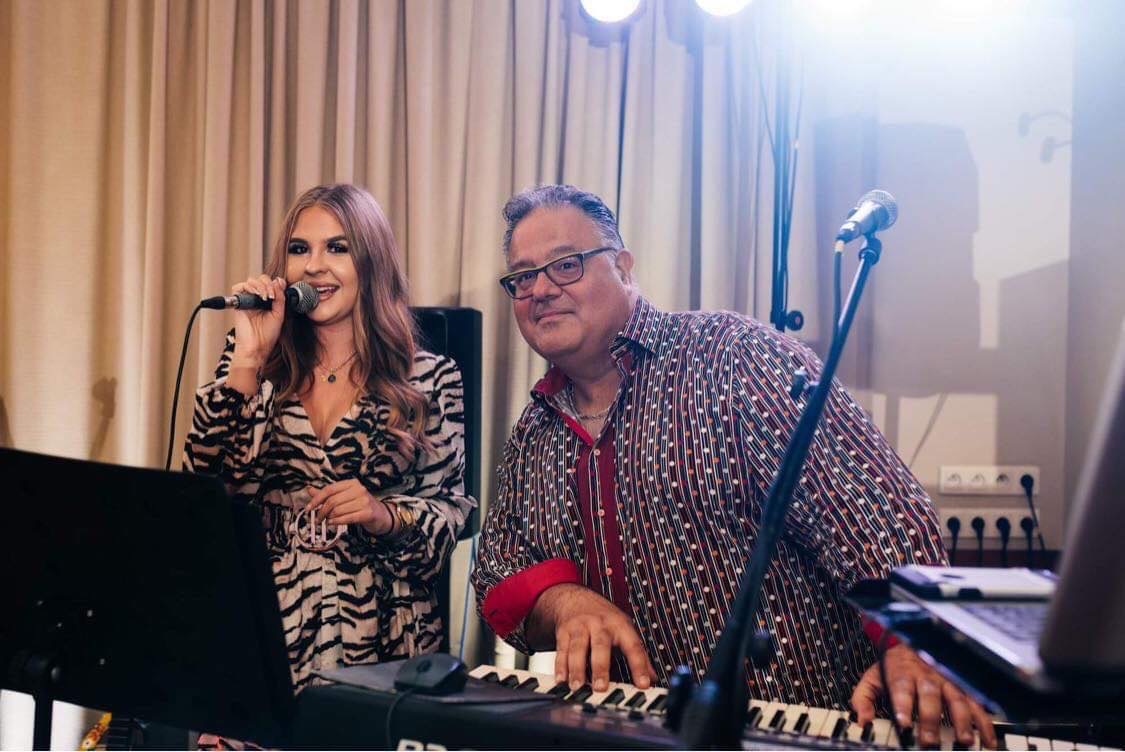 Wokalistka w sukience ze wzorem zebry śpiewa do mikrofonu, obok pianista w koszuli w kropki gra na keyboardzie, za nimi kremowe zasłony i reflektory.