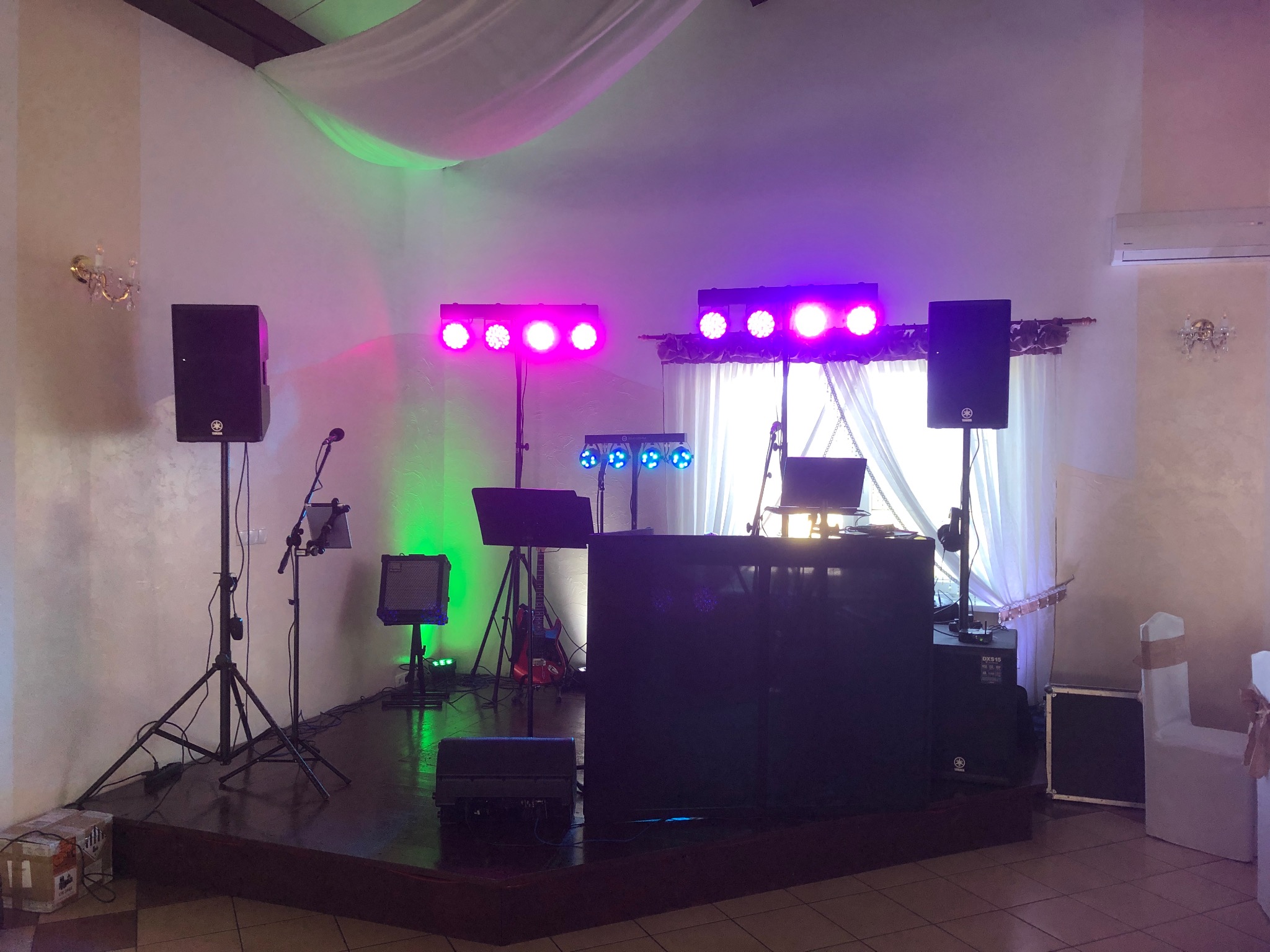 Scena z profesjonalnym sprzętem nagłośnieniowym i oświetleniowym gotowa do imprezy, widoczne kolumny Yamaha, reflektory LED w kolorach różowym, zielonym i niebieskim, mikser DJ-ski oraz gitara...