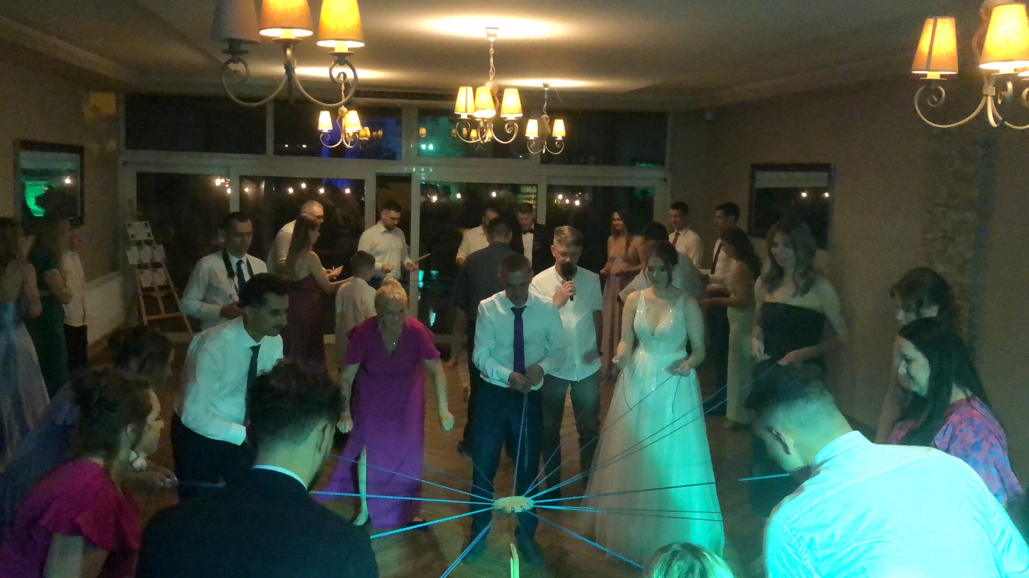 Goście weselni uczestniczący w zabawie z niebieskimi sznurkami, w tle para młoda i konferansjer z mikrofonem, oświetlenie sali z żyrandolami