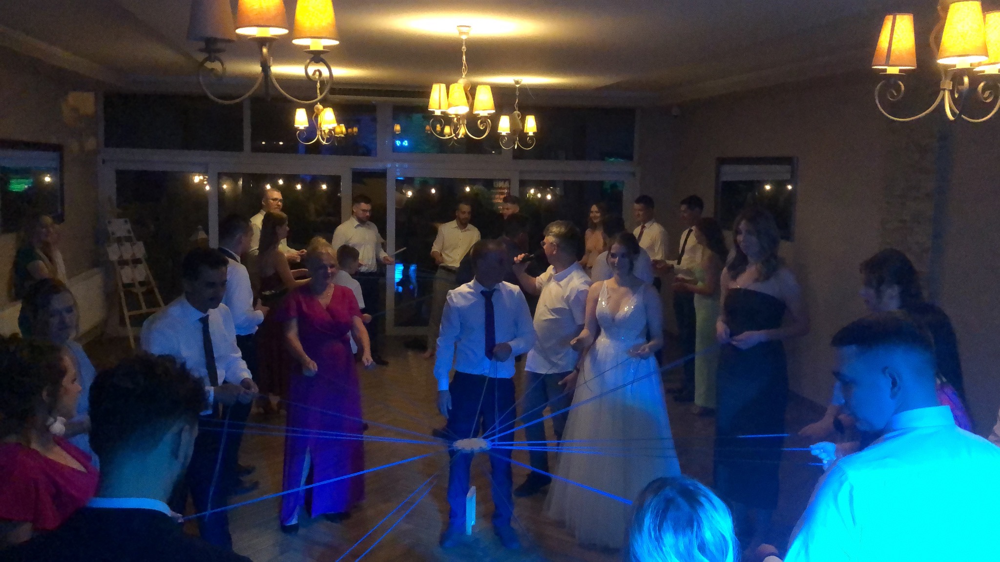 Grupa osób uczestniczy w zabawie z niebieskimi sznurkami podczas uroczystości w sali z żyrandolami.