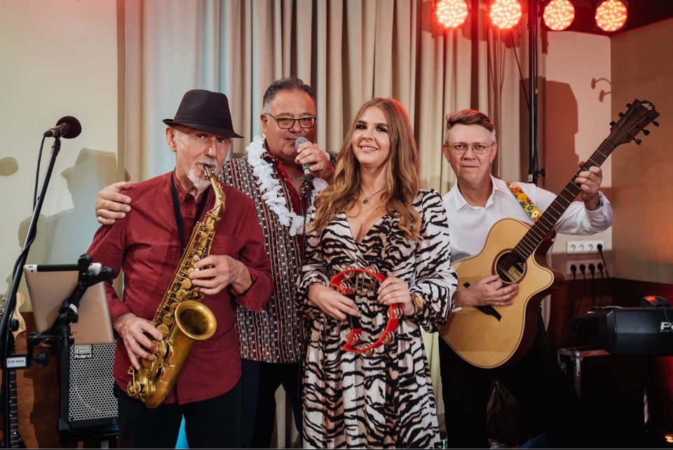 Zespół muzyczny: saksofonista w kapeluszu, wokalista z mikrofonem w hawajskim naszyjniku, kobieta z tamburynem w sukience w zebrę i gitarzysta w białej koszuli, oświetlenie sceniczne w tle.