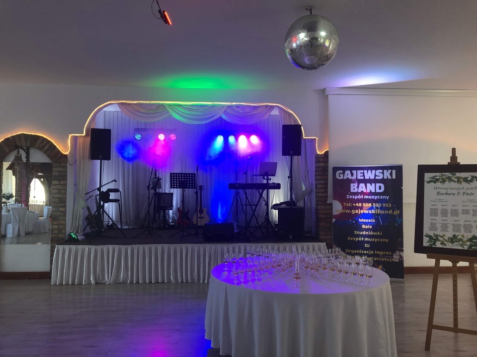 Scena z nagłośnieniem i oświetleniem przygotowana na imprezę, z widocznym banerem zespołu muzycznego 'Gajewski Band' i stołem z kieliszkami w tle.