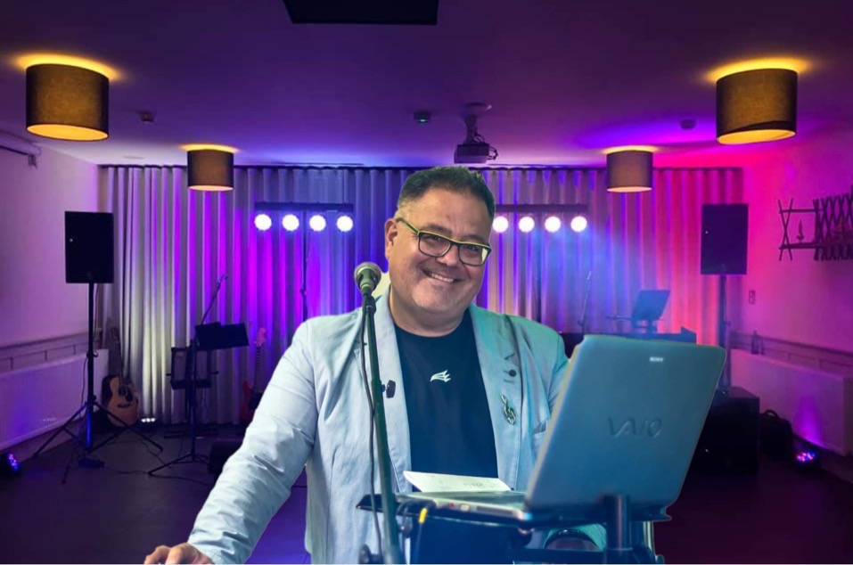 Uśmiechnięty mężczyzna w okularach i jasnej marynarce, stoi za mikrofonem i laptopem marki Sony, na tle sceny z nagłośnieniem, oświetleniem i instrumentami muzycznymi.