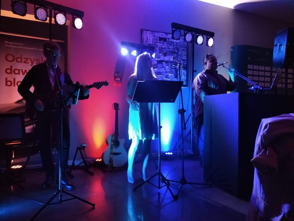 Zespół muzyczny podczas występu na scenie oświetlonej kolorowymi reflektorami, gitarzysta, wokalistka i klawiszowiec, gitara akustyczna oparta o statyw w tle.