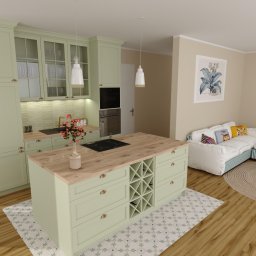 Pracownia Ładne Rzeczy Karolina Szczepanik - Jasna, pistacjowa kuchnia z wyspą i drewnianym blatem, widoczna z perspektywy salonu z sofą i okrągłym dywanem. Minimalistyczne lampy wiszące nad wyspą.