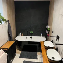 Nowoczesna łazienka z wolnostojącą czarną wanną, dwiema umywalkami nablatowymi na drewnianym blacie i czarną armaturą na tle ściany z czarnych płytek.