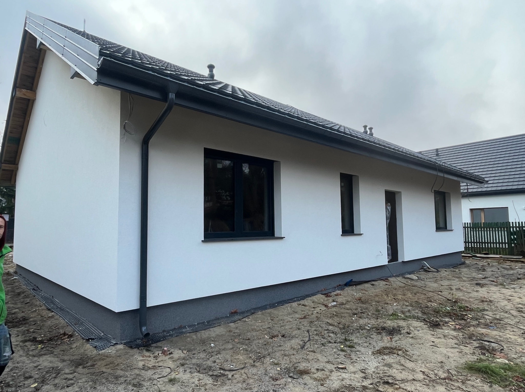 Świeżo otynkowany, parterowy dom jednorodzinny z ciemną stolarką okienną i dachem krytym czarną dachówką, widok z boku na tle pochmurnego nieba.