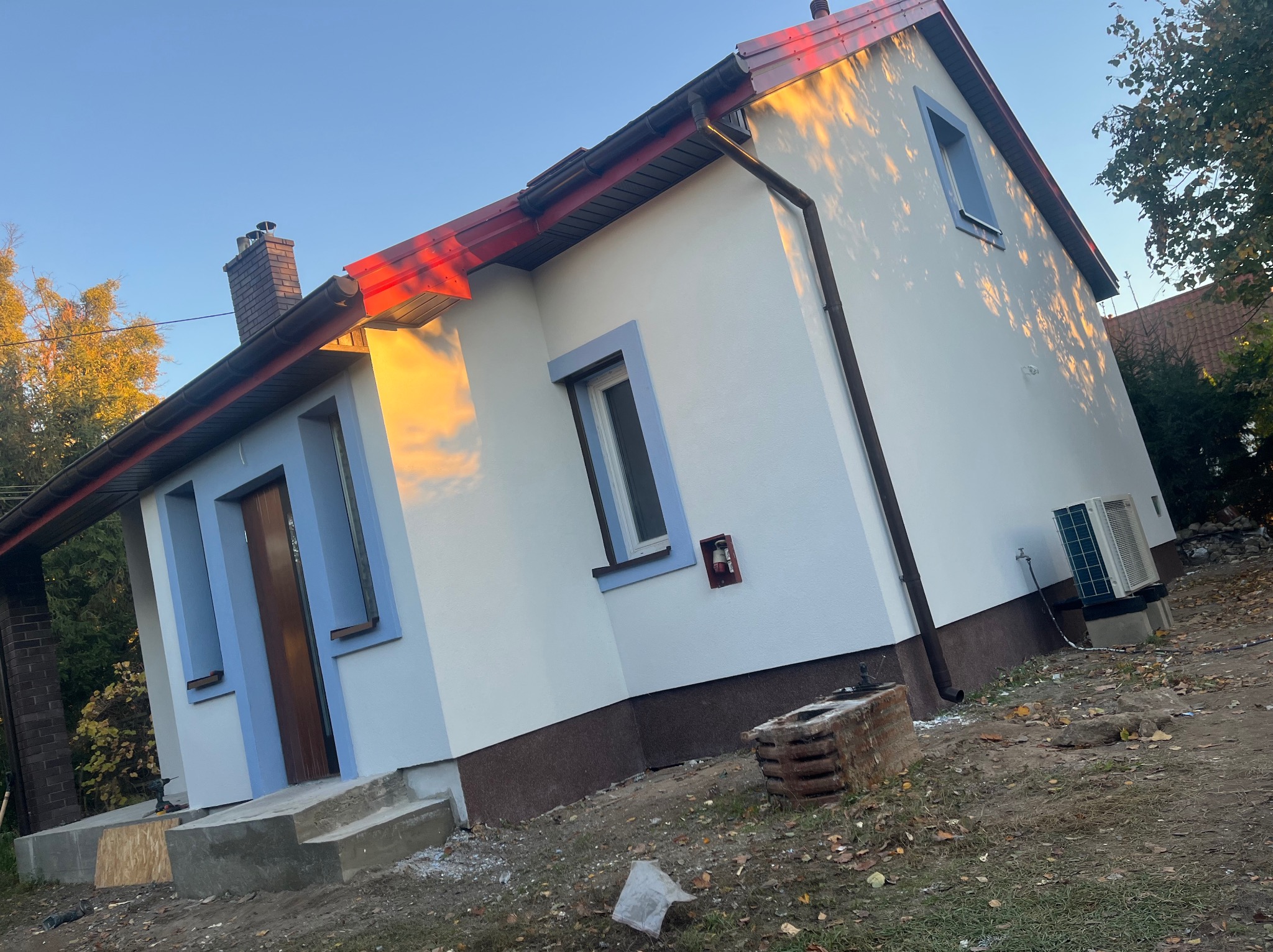 Świeżo otynkowany dom jednorodzinny z niebieskimi ramami okiennymi, widoczne elementy elewacji w kolorze brązowym, czerwony dach i komin z cegły, w tle drzewa i błękitne niebo, na ziemi gruz...