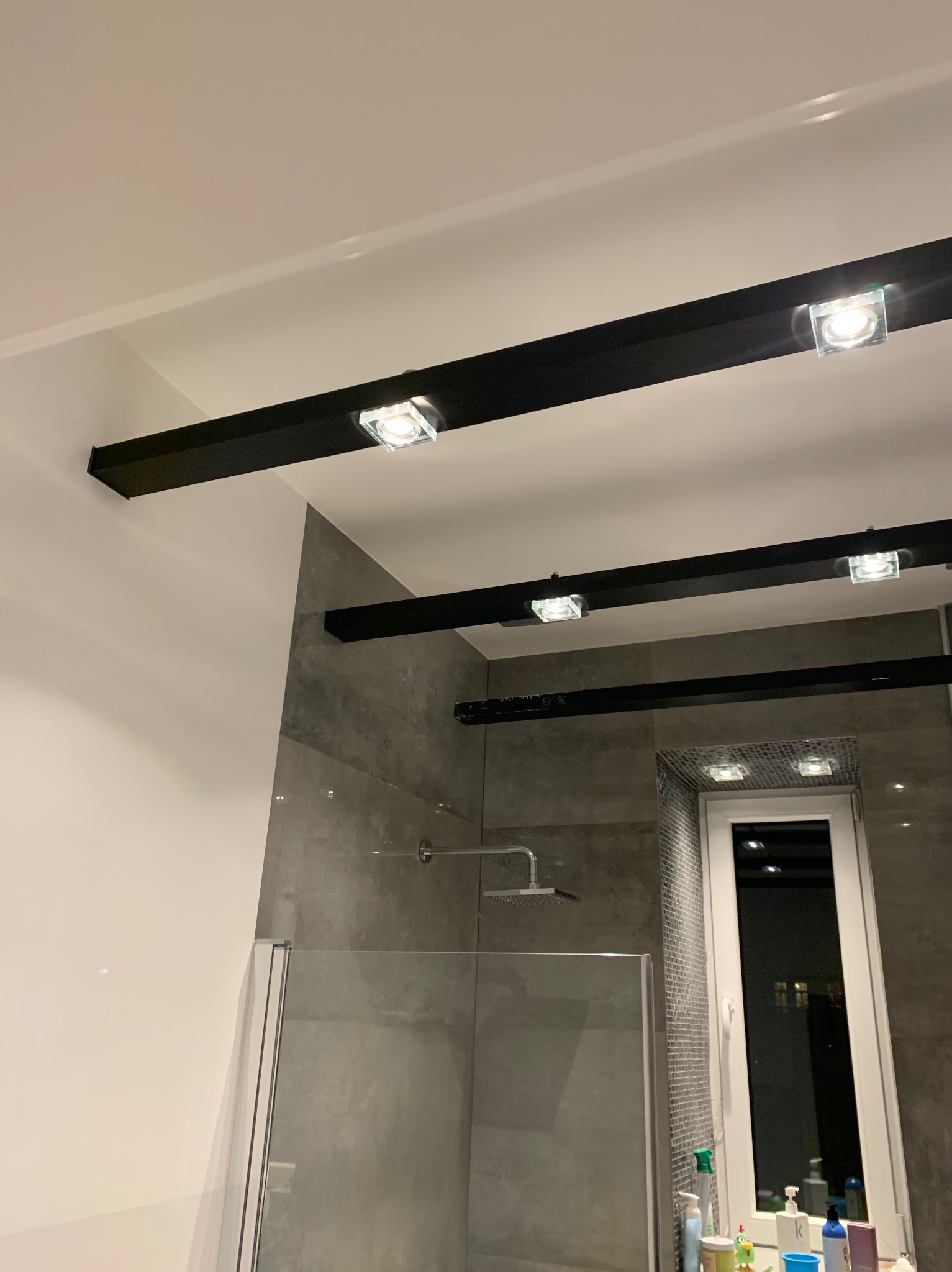 Nowoczesna łazienka z szarą glazurą, kabiną prysznicową i oryginalnym oświetleniem sufitowym na czarnych belkach z kwadratowymi lampami.