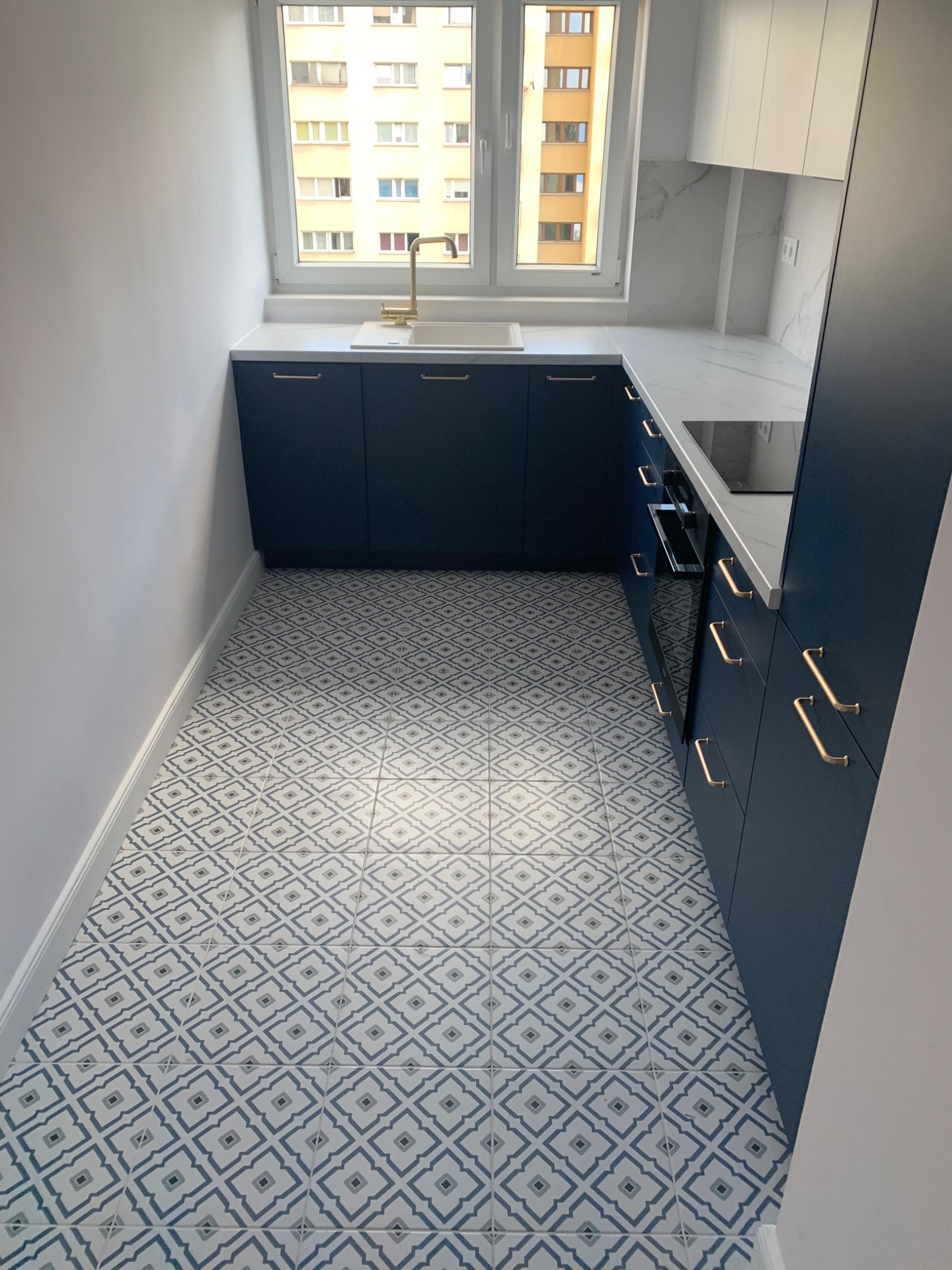Wąska kuchnia z granatowymi szafkami, złotymi uchwytami i baterią, białym blatem imitującym marmur oraz wzorzystą podłogą w odcieniach szarości i błękitu.