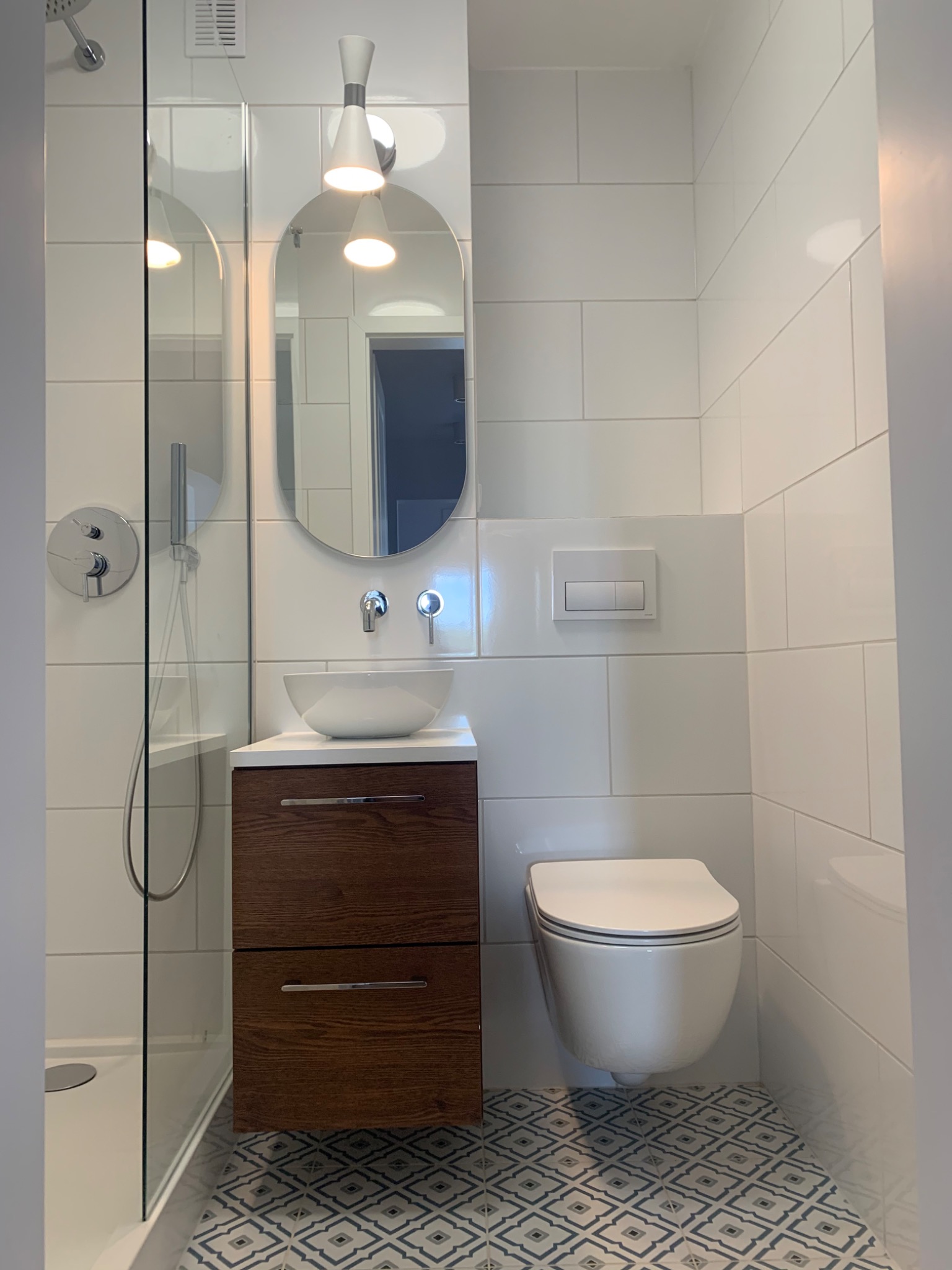 Nowoczesna łazienka z białą ceramiką, umywalką nablatową na drewnianej szafce, wiszącą toaletą i kabiną prysznicową za szkłem, wykończona białymi płytkami i wzorzystą podłogą.