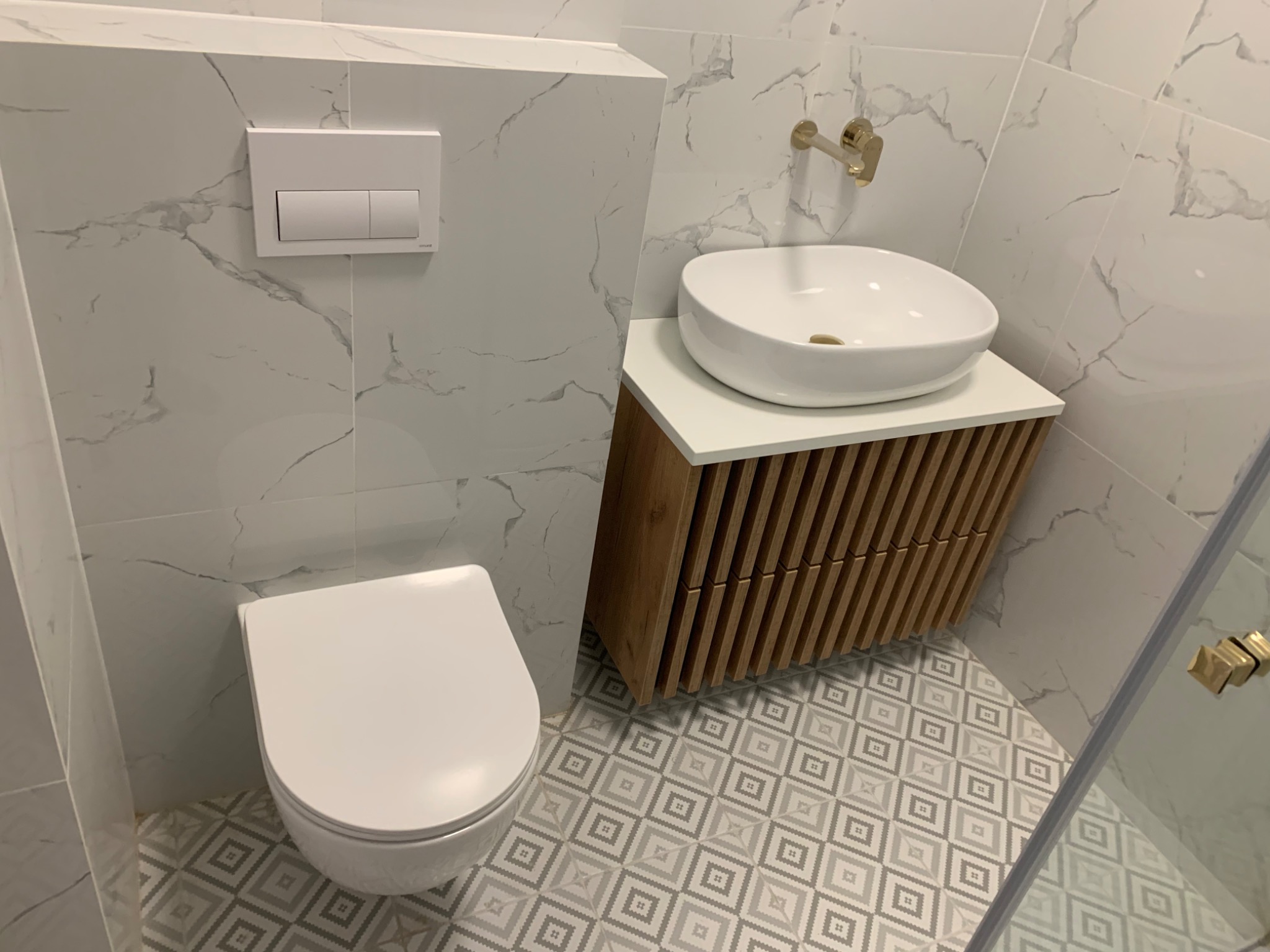 Nowoczesna łazienka z marmurowymi płytkami, wiszącą miską WC, umywalką nablatową na szafce z drewnianymi lamelami i złotą armaturą.